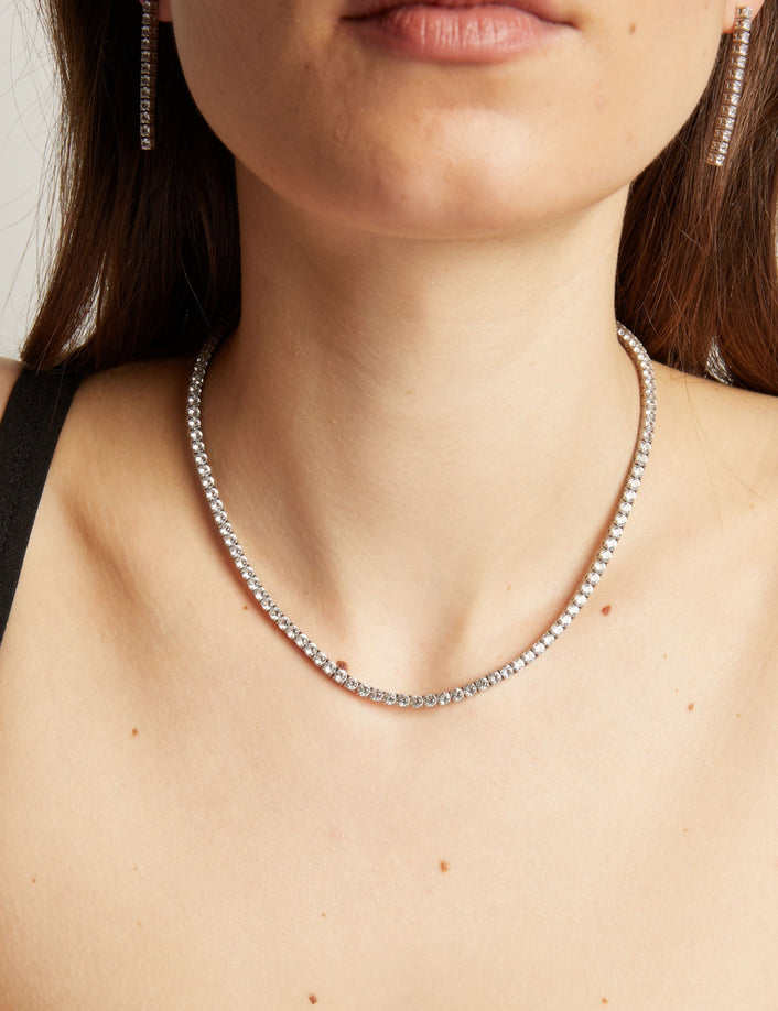 Close up of a woman wearing a tennis necklace and earrings against a neutral background.