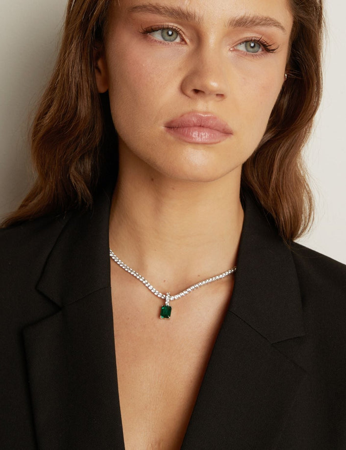 Woman wearing a black blazer with a tennis necklace with a emerald gemstone pendant.