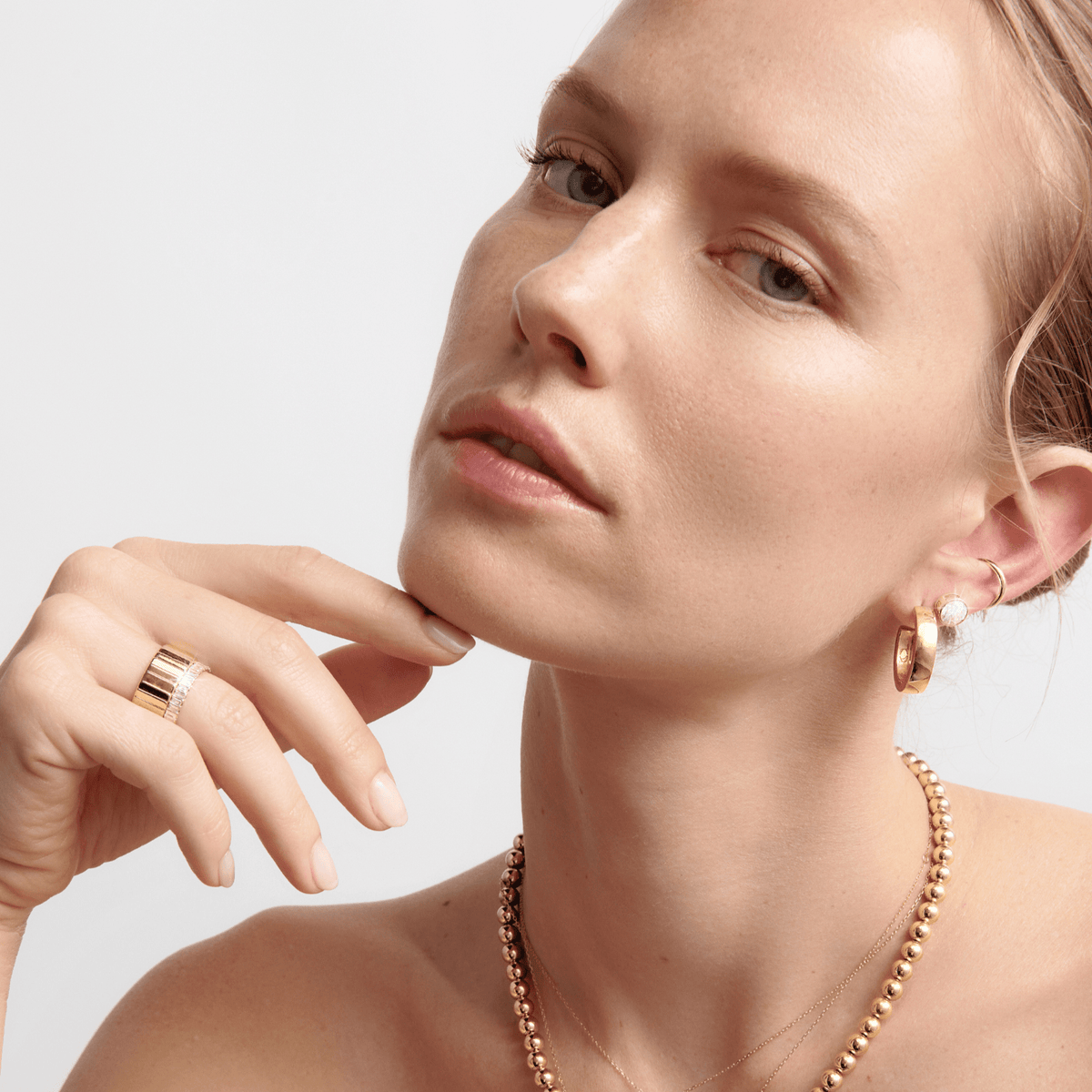 Lifestyle Model Close-up of a woman looking away from the camera with her hand resting gently near her chin against a light background. She is wearing a gold beaded necklace, a thick gold and diamond stacked ring, and multiple earrings, including a thick gold hoop, a pearl stud, and a small gold cuff.