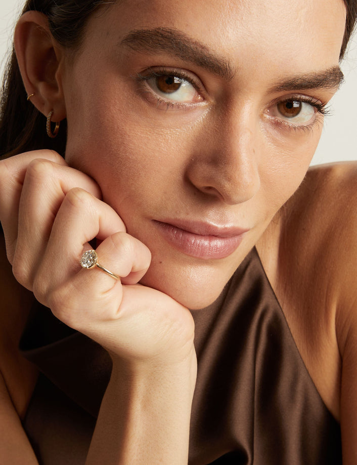 Model. A close-up lifestyle shot of a woman wearing a delicate gold ring with a round diamond. 