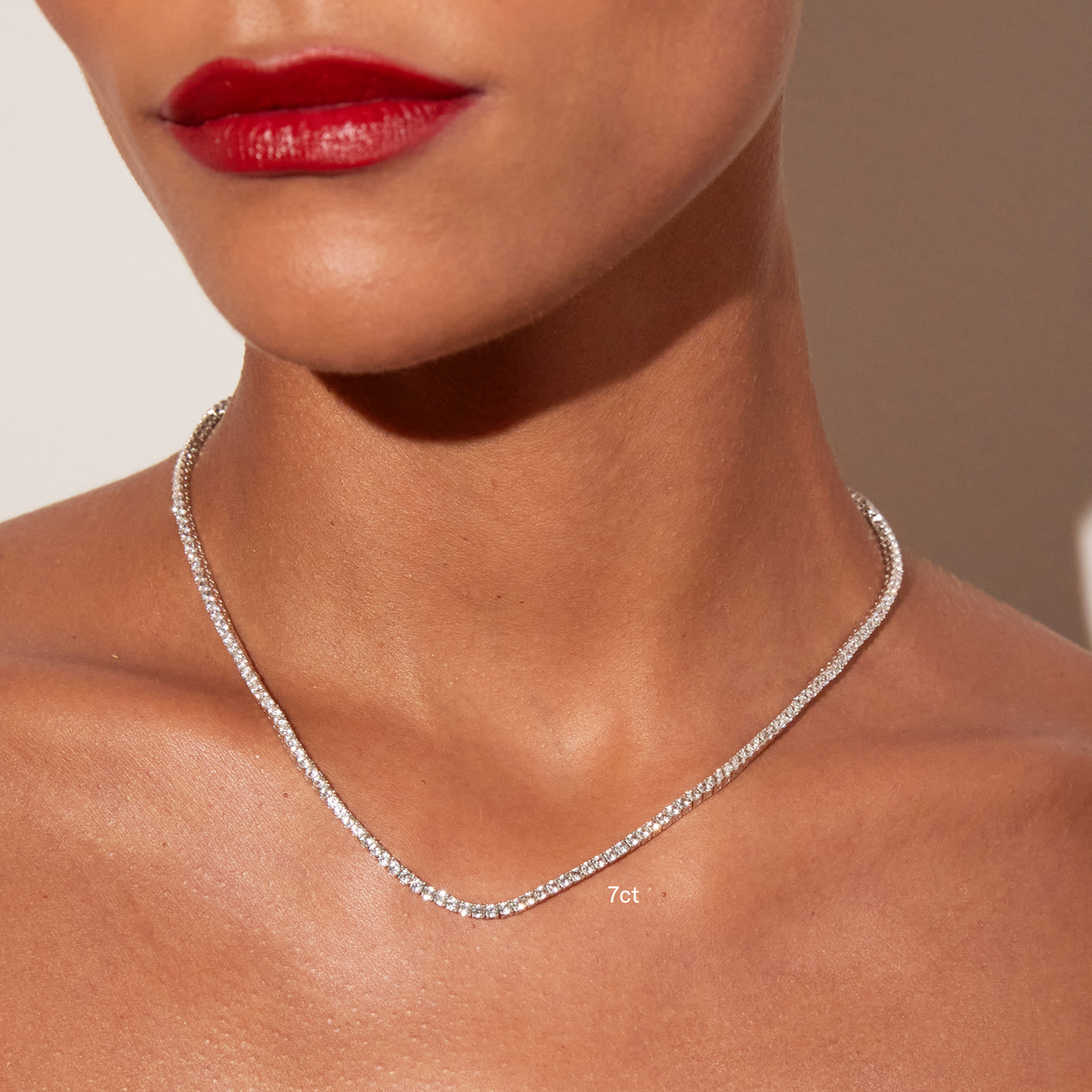 Model. Close-up of a woman's neck and lower face featuring bright red lipstick, wearing a single white sapphire tennis necklace with a '7ct' text overlay, against a warm neutral background.