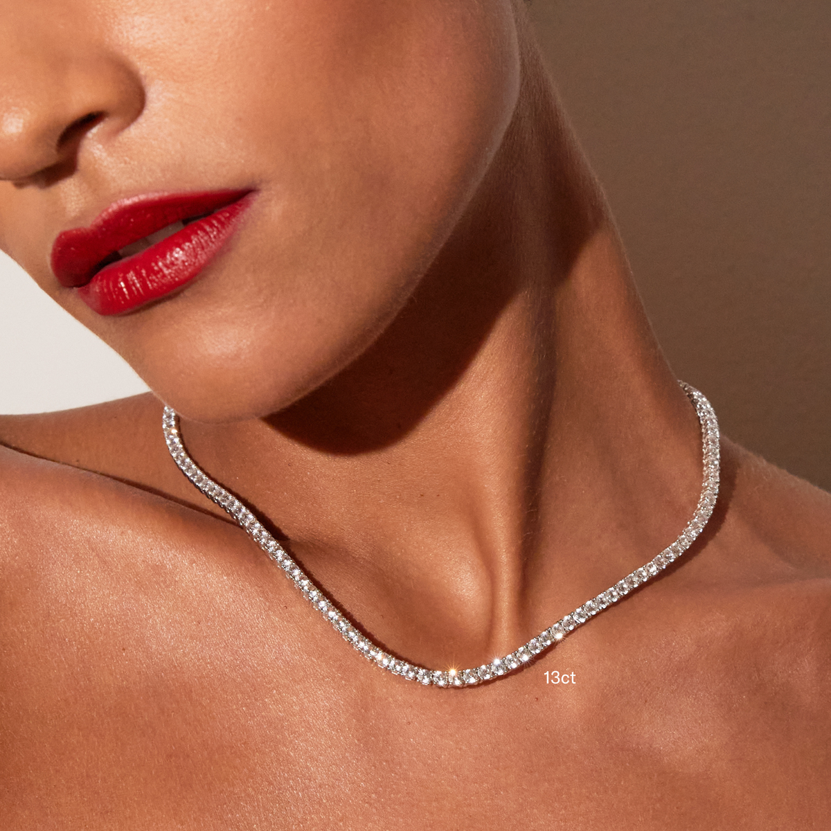 Model. Close-up of a woman's neck and lower face featuring bright red lipstick, wearing a single white sapphire tennis necklace with a '13ct' text overlay, against a warm neutral background