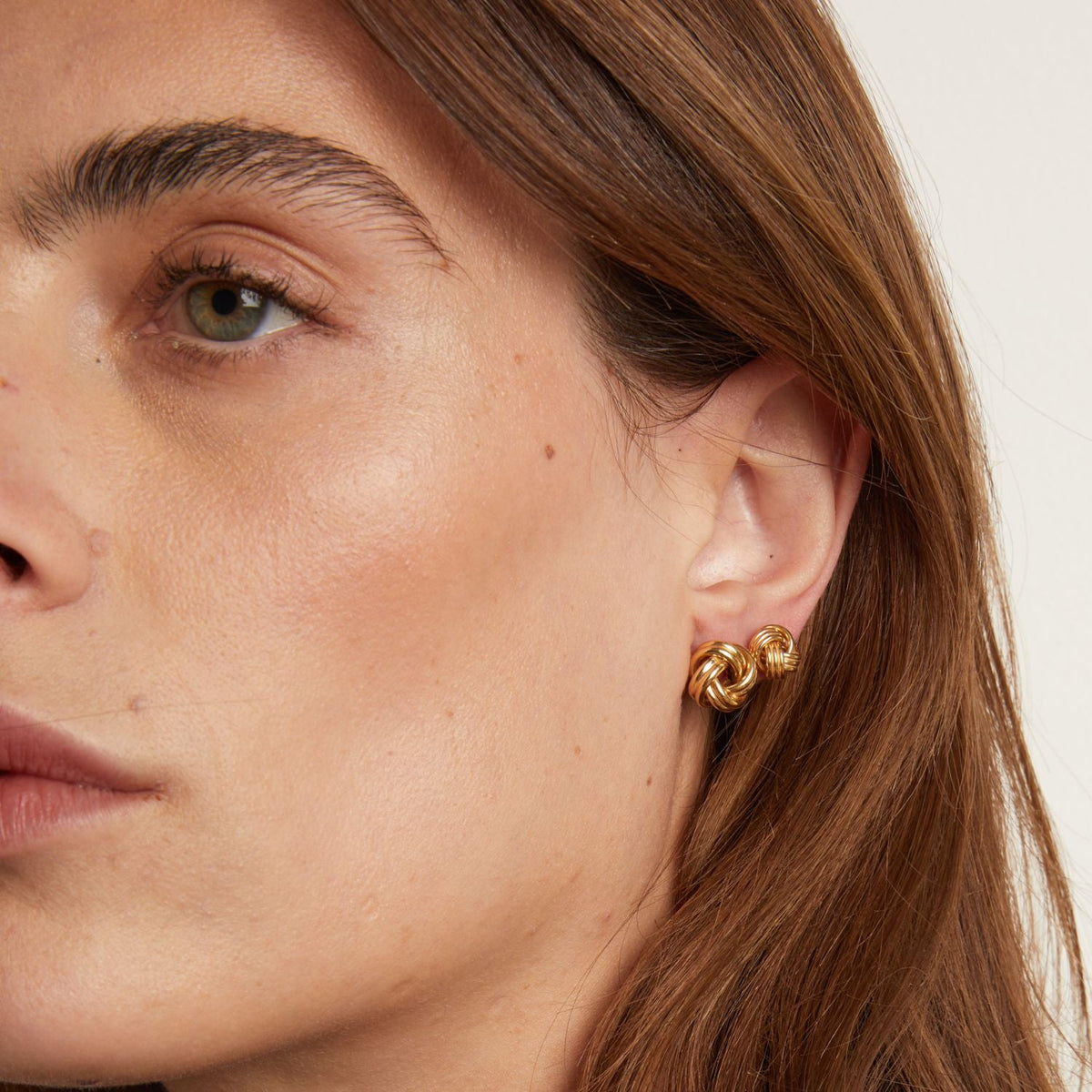 Model. Close-up of a person wearing gold hoop earrings with a neutral background