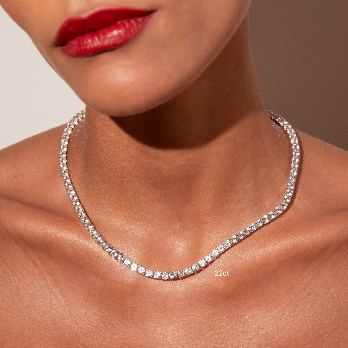 Model. Close-up of a woman's neck and lower face featuring bright red lipstick, wearing a single white sapphire tennis necklace with a '22ct' text overlay, against a warm neutral background.
