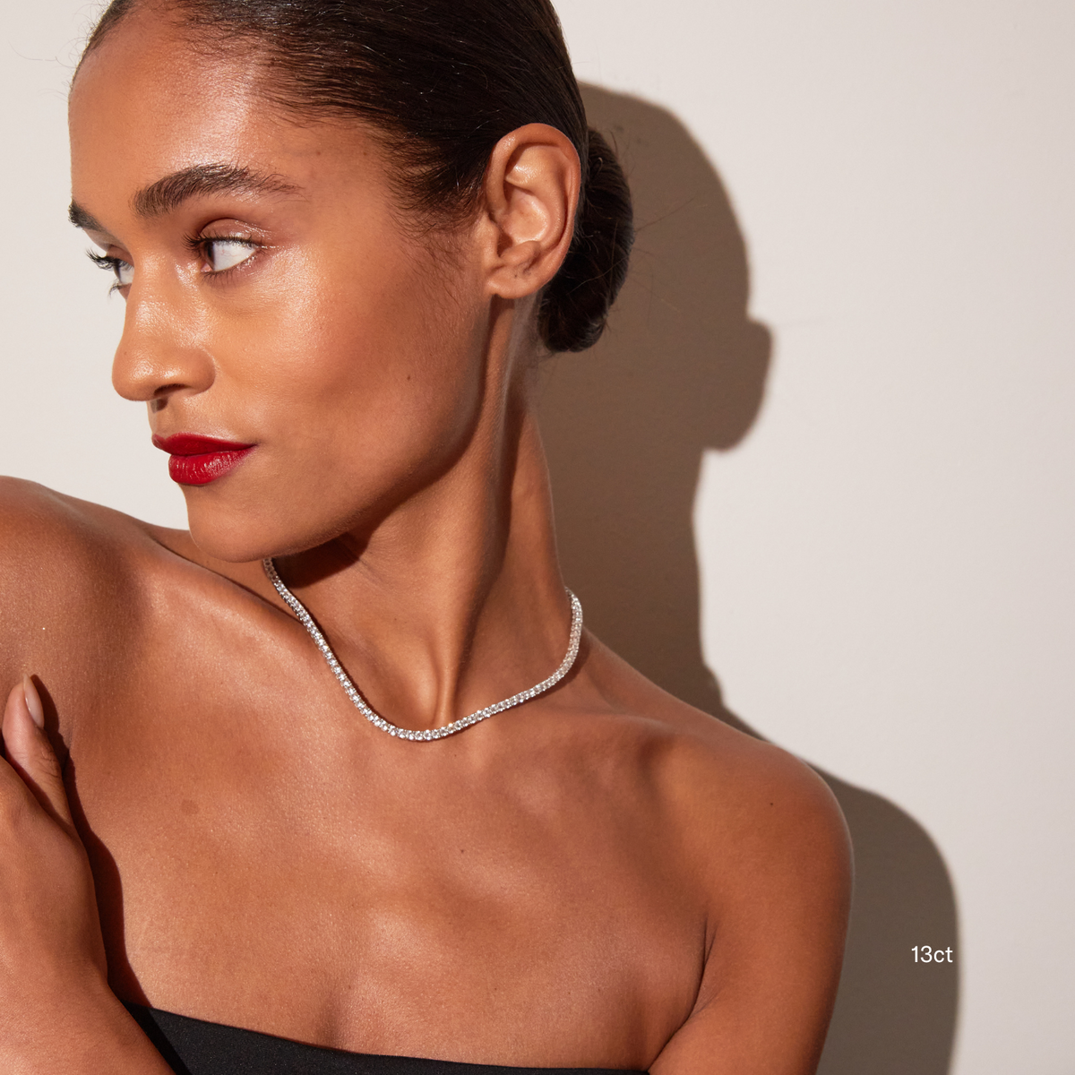 Model. Portrait of a woman looking off-camera, wearing a black strapless top and bright red lipstick. She is wearing a single white sapphire tennis necklace against a warm neutral background with a distinct shadow, with a '13ct' text overlay visible near her shoulder