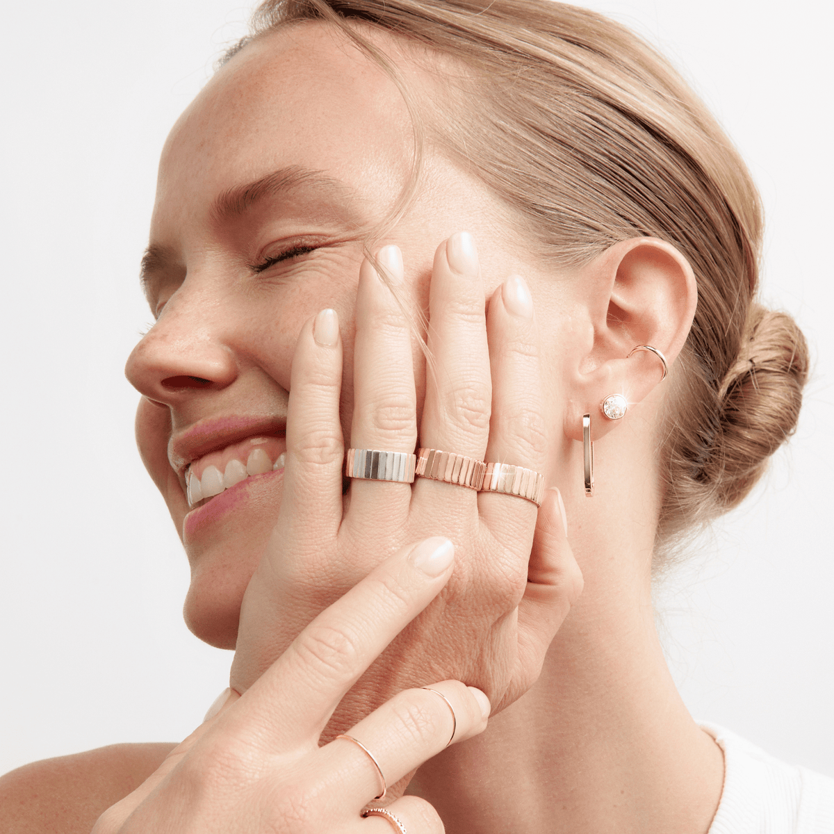 Lifestyle Model Close-up of a smiling woman's profile with her eyes closed and her hand gently resting near her face. She is wearing a stack of gold rings, including a thick smooth band and a textured ribbed band, along with multiple earrings: a round bezel-set diamond stud, a small gold hoop, and a long rectangular drop earring.