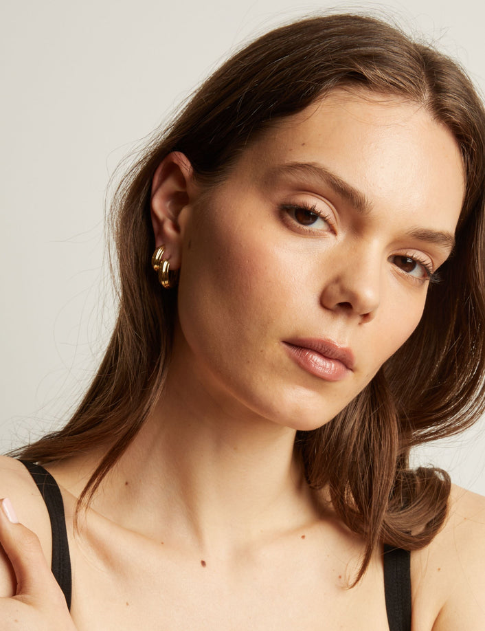Model. Close up of a person wearing gold hoop earrings with a neutral background