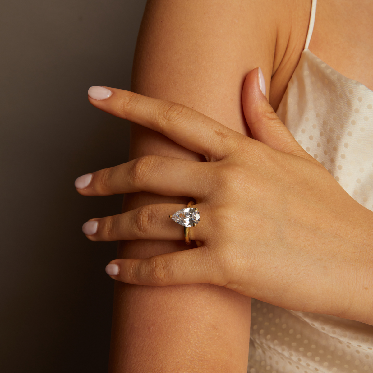 Model. Hand wearing a gold ring with a pear-shaped diamond on a neutral background