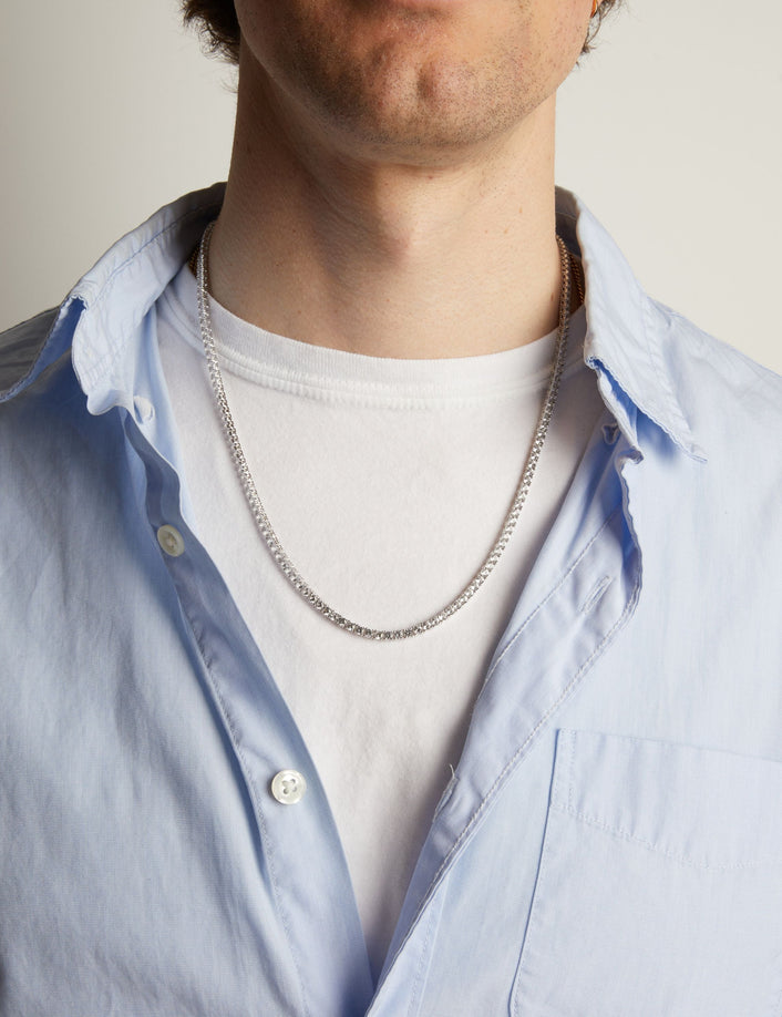 A close-up of a man wearing a tennis necklace over a white t-shirt and a light blue button-down shirt.