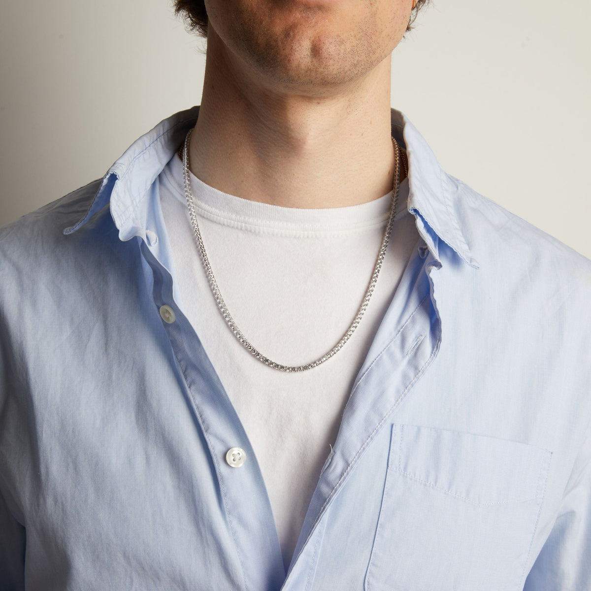 A close-up of a man wearing a tennis necklace over a white t-shirt and a light blue button-down shirt.
