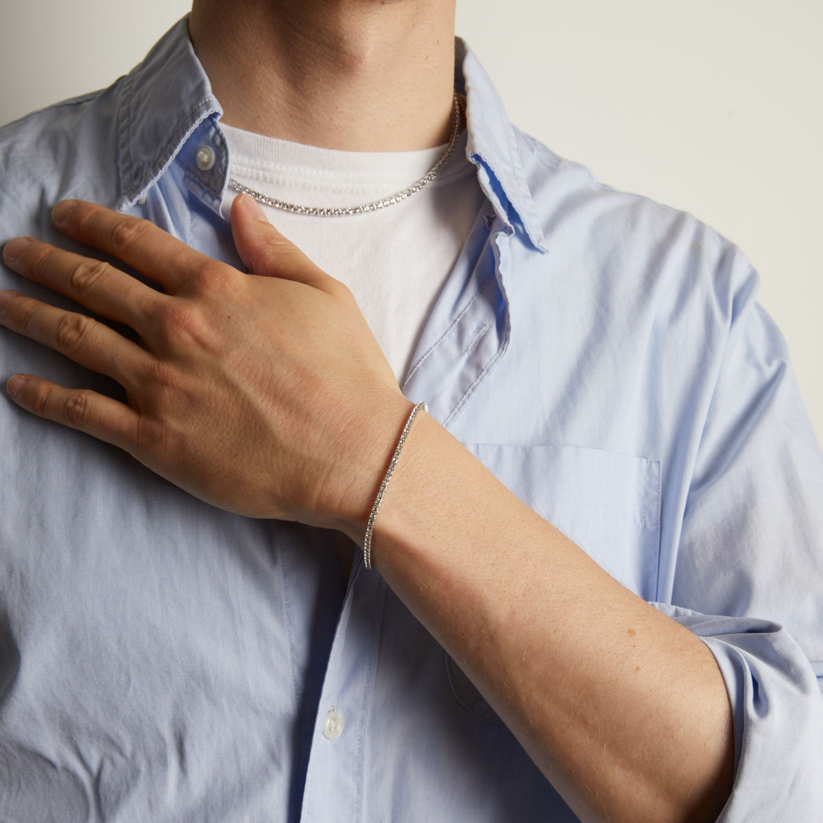 A close-up of a man's wrist wearing a delicate tennis bracelet, shown against a light blue button-down shirt.