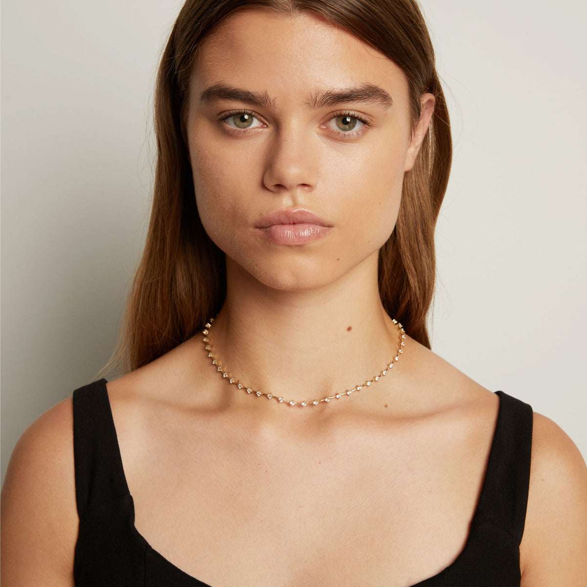 Model. A close-up of a model wearing a delicate gold chain necklace with small clear stones, set against a soft neutral background.