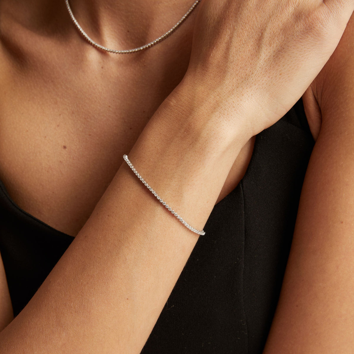 Model. A close-up lifestyle shot of a woman wearing a delicate silver tennis necklace and a matching silver tennis bracelet, paired with a black top.