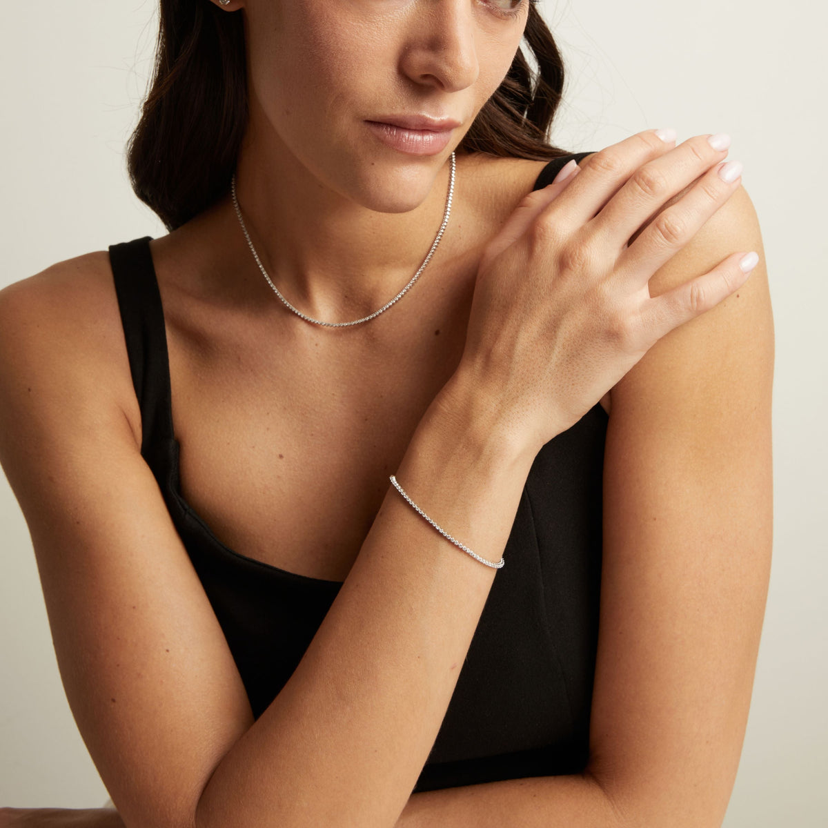 Model. A close-up lifestyle shot of a woman wearing a delicate silver tennis necklace and a matching silver tennis bracelet, paired with a black top.