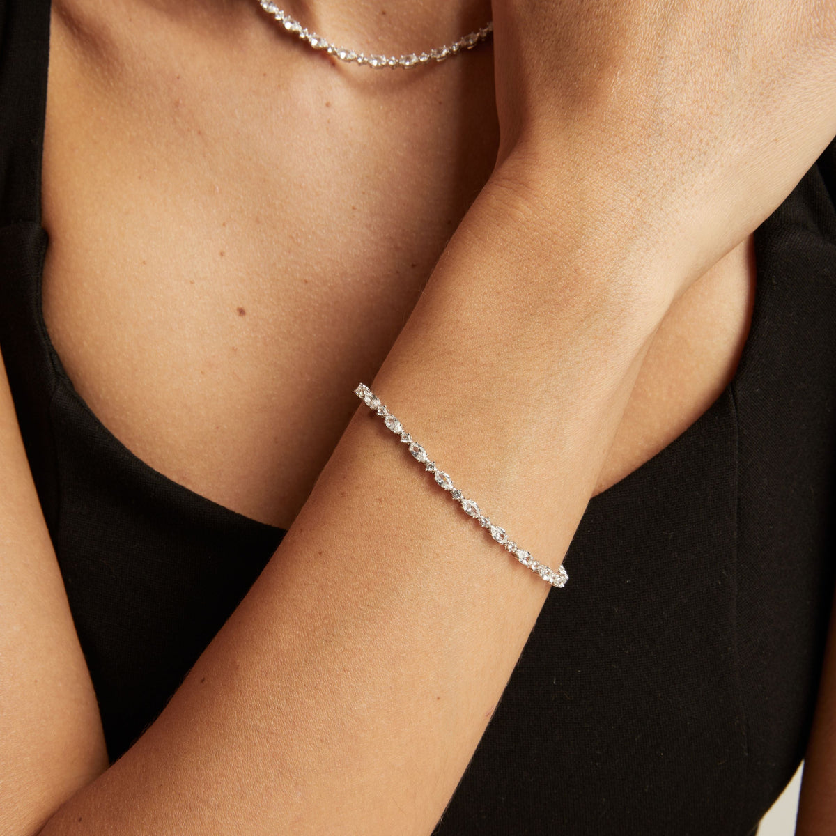 Model. A close-up of a woman's wrist wearing a delicate silver tennis bracelet, with a matching silver chain necklace partially visible at the top against a black top.