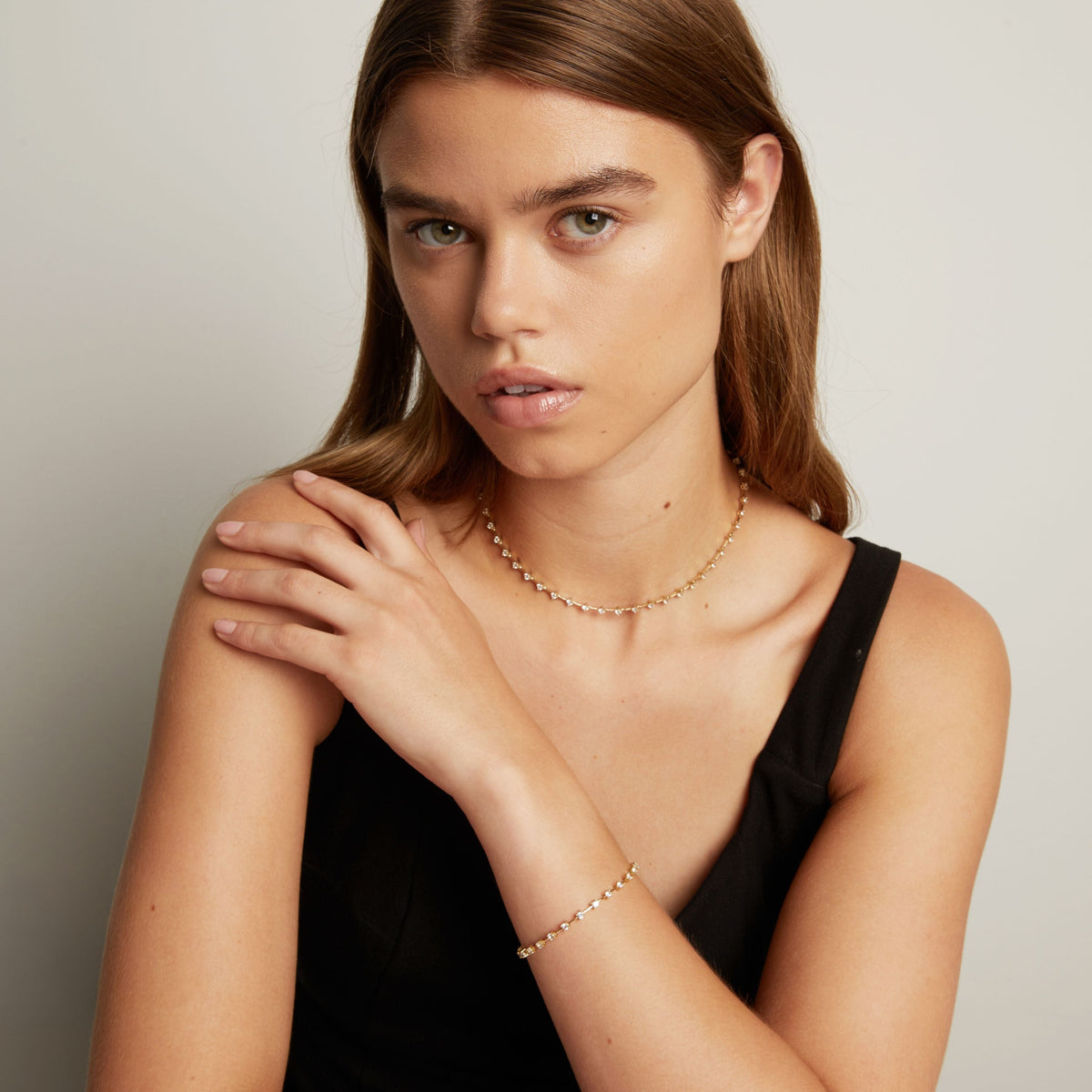 Model. A close-up of a model wearing a delicate gold chain necklace with small clear stones, set against a soft neutral background.