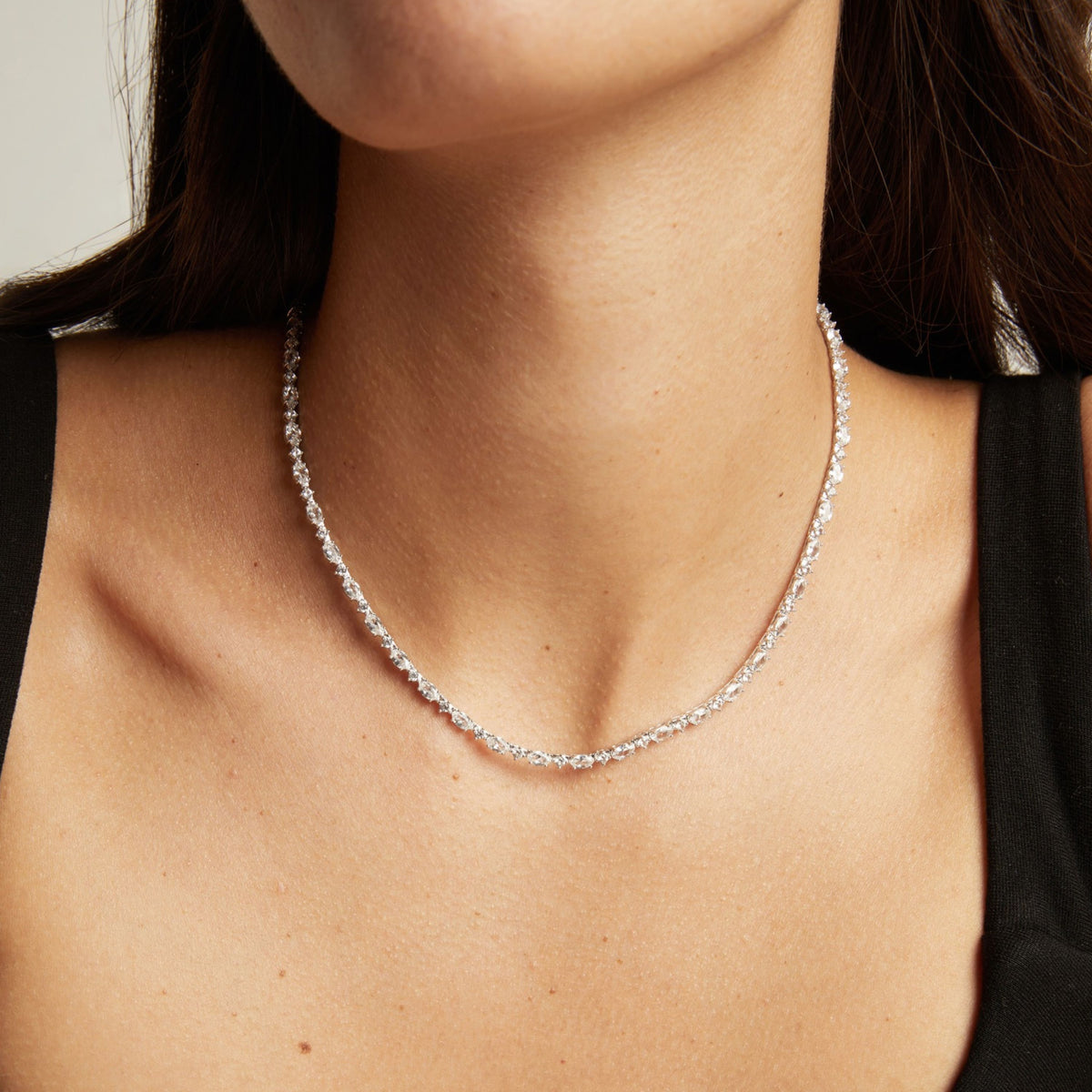 Model. A close-up of a delicate silver tennis necklace with clear stones worn on a woman's neck. Styled with a black top against a light neutral background.