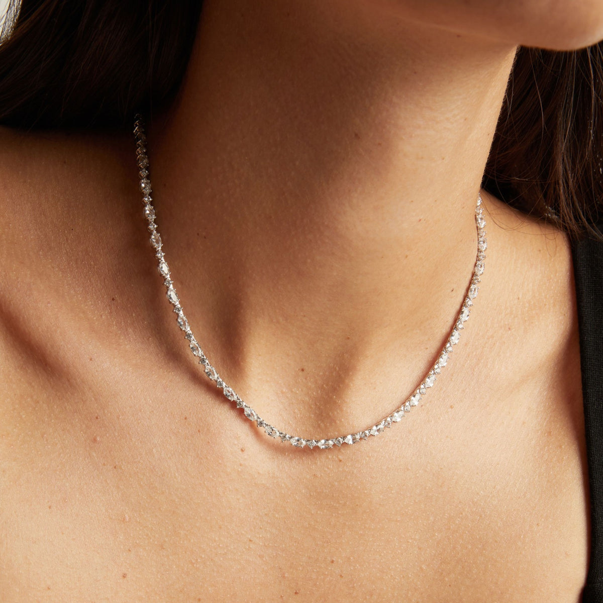Model. A close-up of a delicate silver tennis necklace with clear stones worn on a woman's neck.