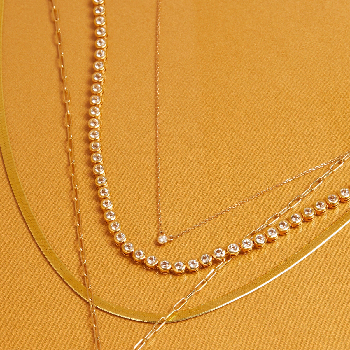Flatlay Model Four layered gold necklaces, including a delicate beaded chain, a chain with a small round gemstone pendant, a diamond tennis necklace, and a paperclip chain, resting on a yellow background.
