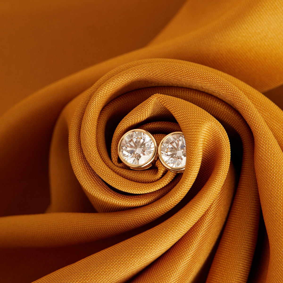 Flatlay Model Close-up of a pair of large, round, bezel-set diamond stud earrings nestled inside the swirling folds of a textured, golden-yellow fabric.
