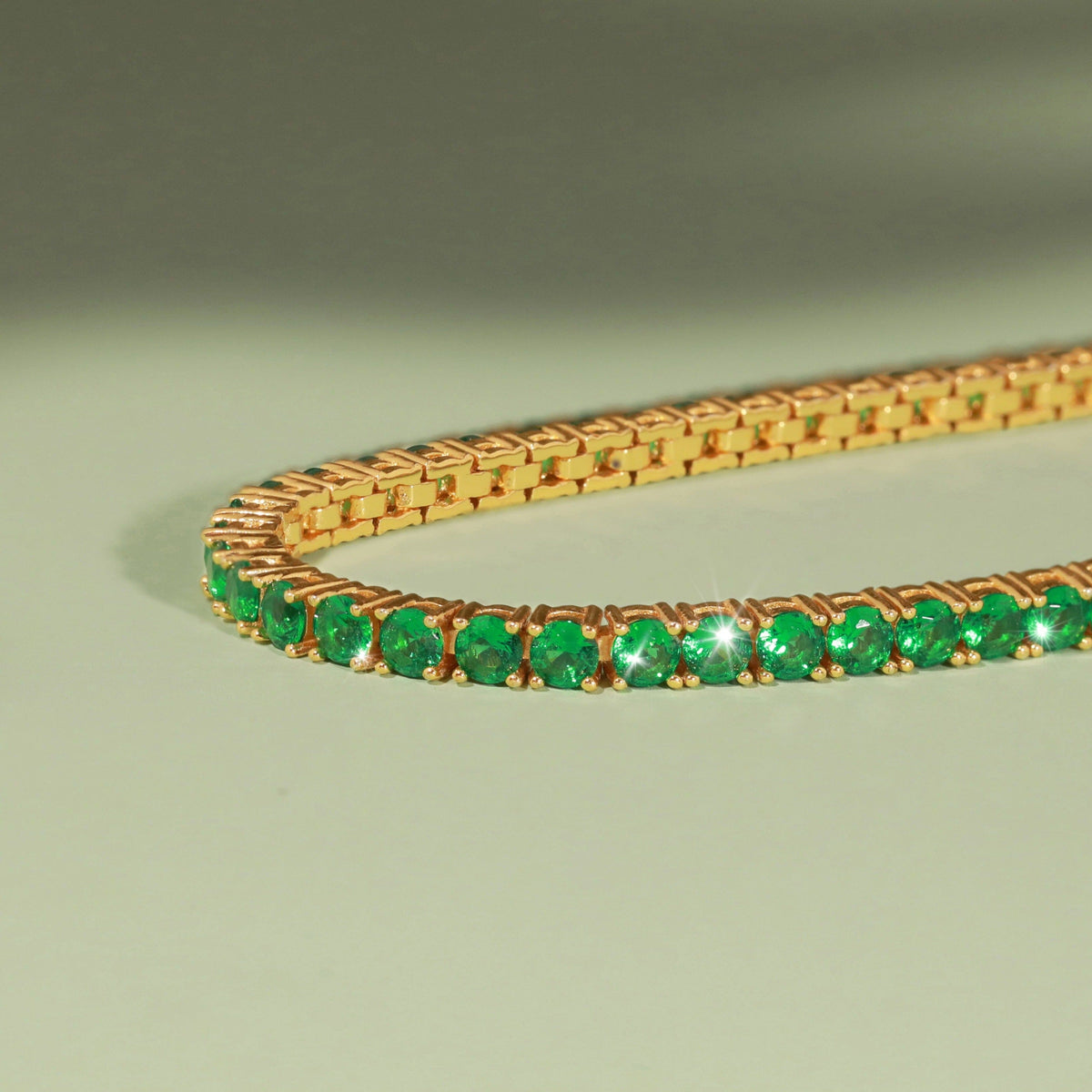 Flatlay Model Close-up of a gold tennis bracelet featuring round green emeralds resting on a light green surface