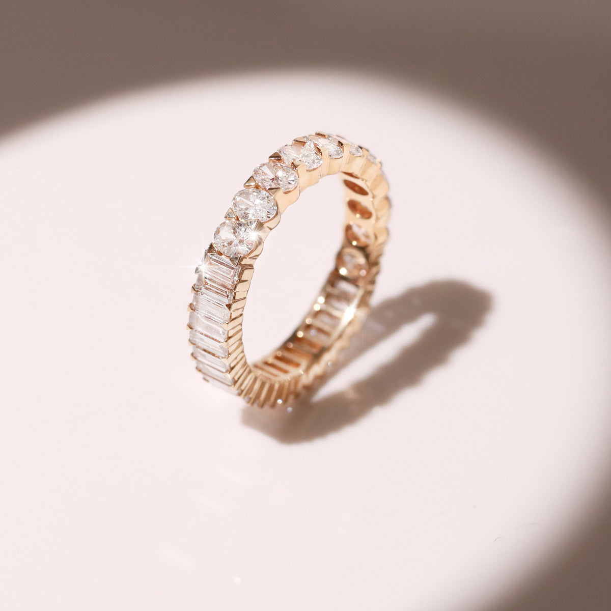 Flatlay Model A close-up of a gold diamond eternity ring standing upright against a plain light background, casting a distinct shadow. The ring features a unique two-in-one design with baguette-cut diamonds on one half and oval-cut diamonds on the other.