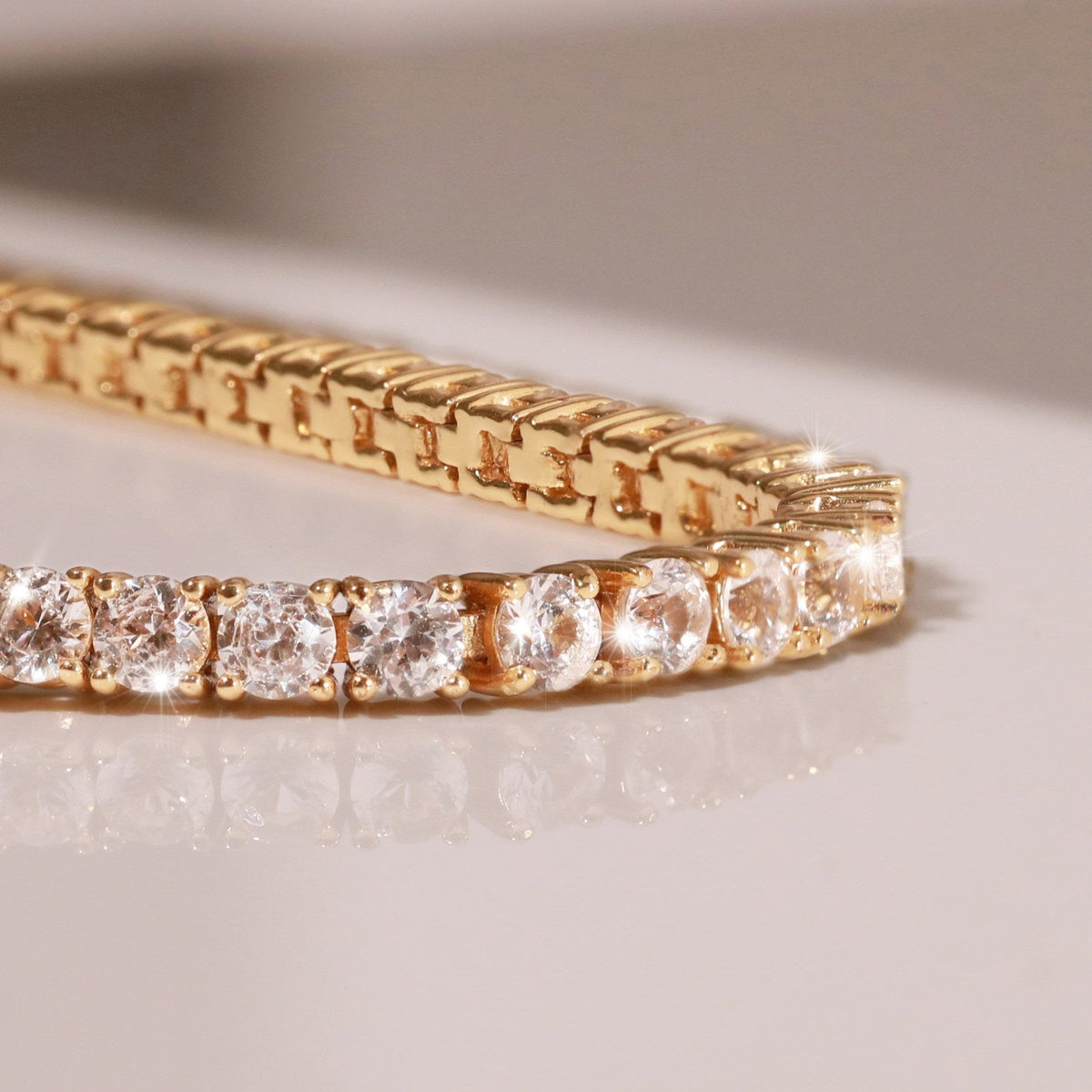 Model.  Close-up of a gold tennis bracelet featuring round white sapphires resting on a light, reflective surface