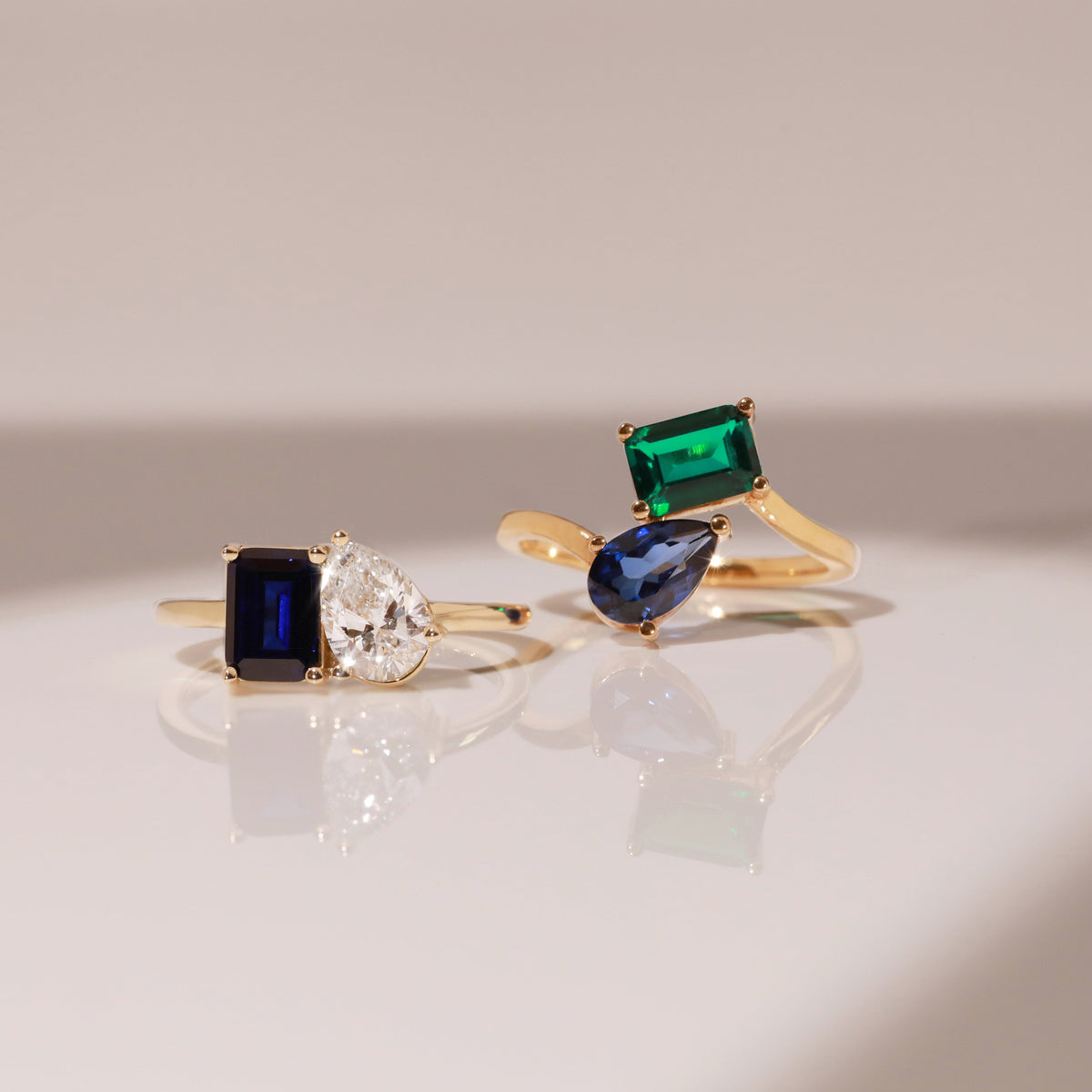 Model Close-up of two gold two-stone rings with different colored gemstones resting on a reflective white surface.