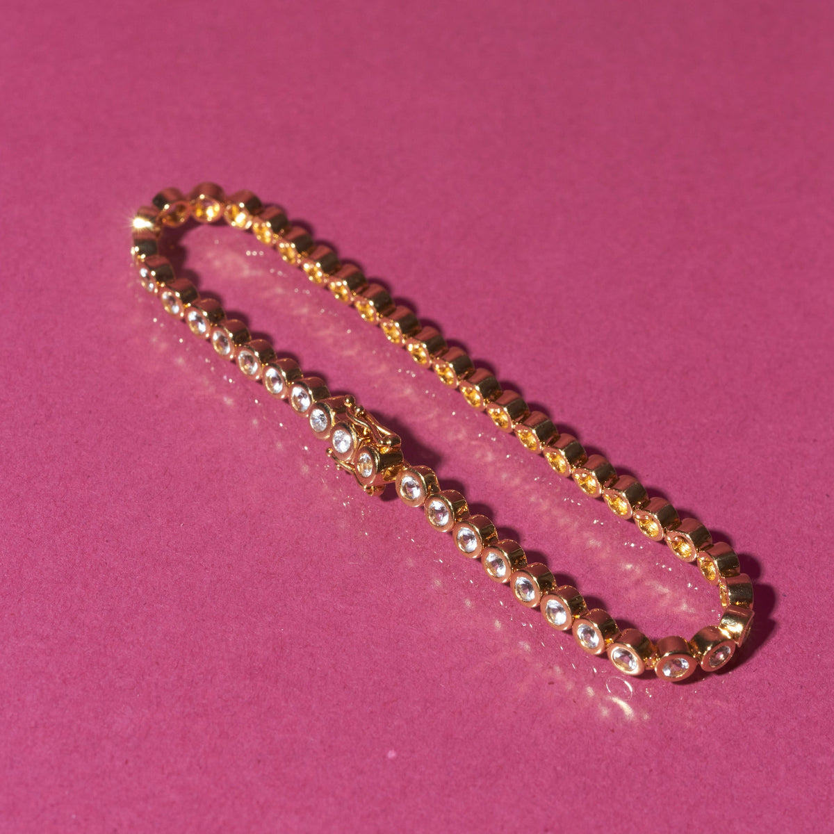 Flatlay Model (Unedited) A single gold diamond bezel tennis bracelet resting diagonally on a bright pink background.