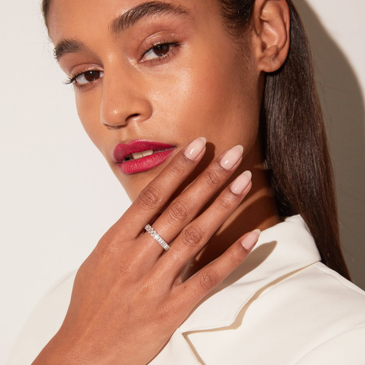 Model Close-up of a woman with her hand resting near her collarbone, wearing a gold diamond eternity ring and a white top, against a warm neutral background