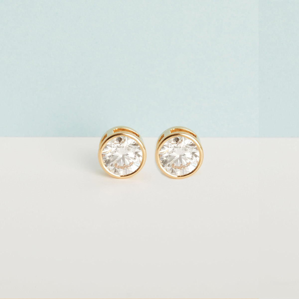 Flatlay Model Close-up of a pair of large, round, bezel-set diamond stud earrings in yellow gold, resting flat against a two-toned background split horizontally with light blue on the top and off-white on the bottom.