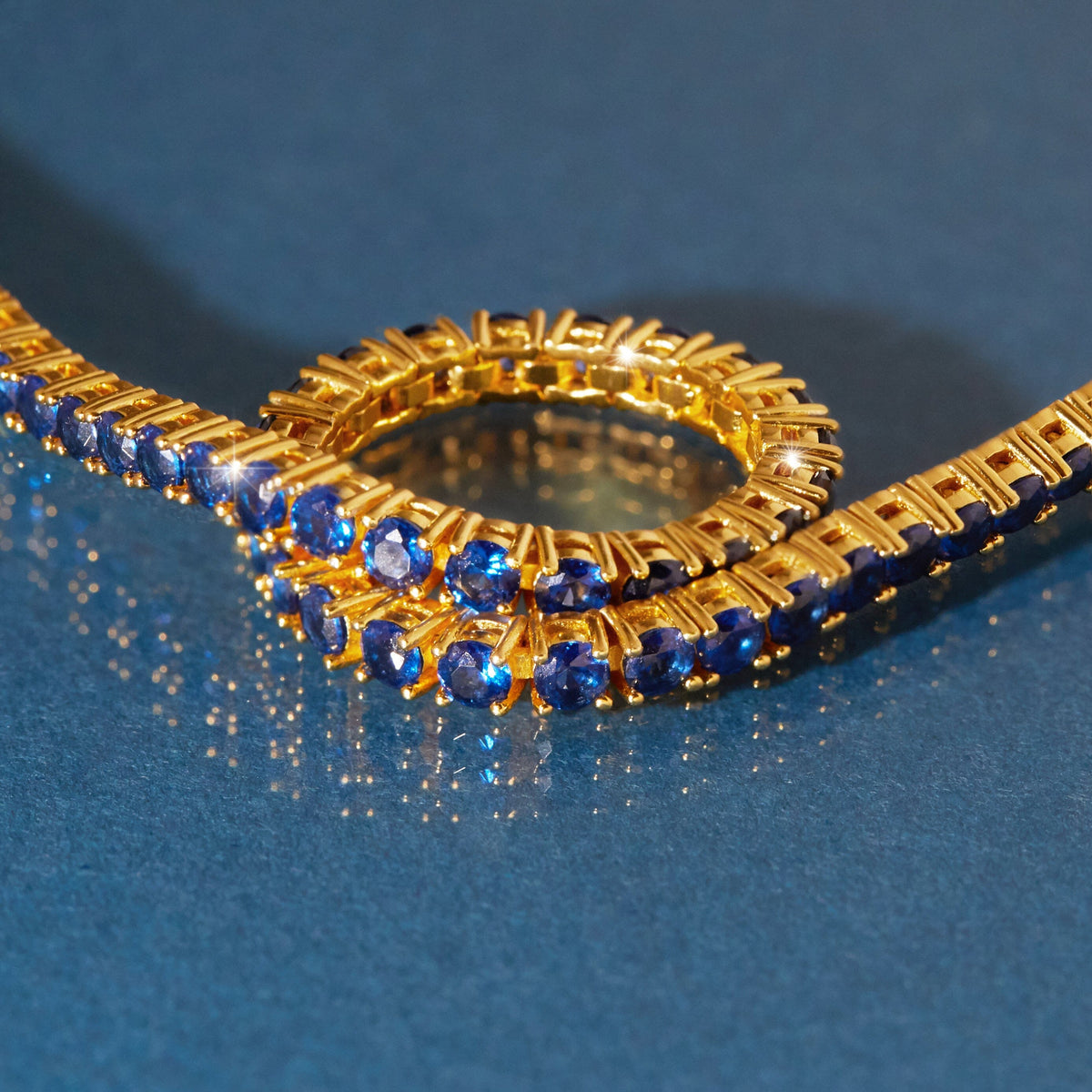 Flatlay Model  Close-up of a gold tennis bracelet featuring round blue sapphires resting on a dark blue reflective surface