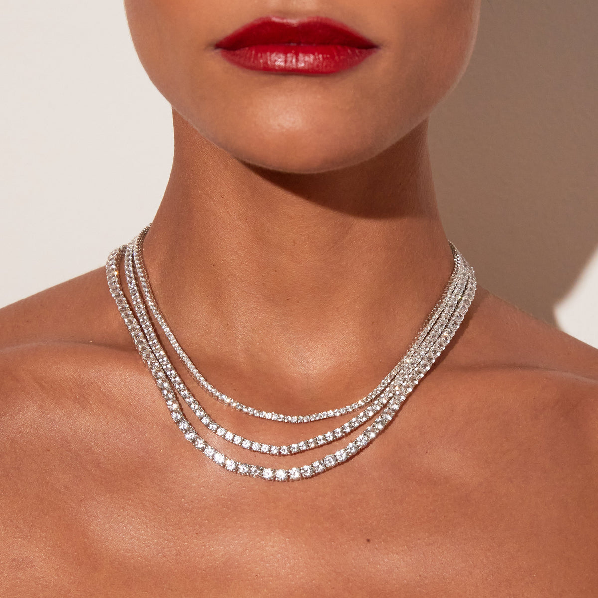Close-up of a woman's neck and lower face featuring bright red lipstick, wearing three layered white sapphire tennis necklaces against a warm neutral background