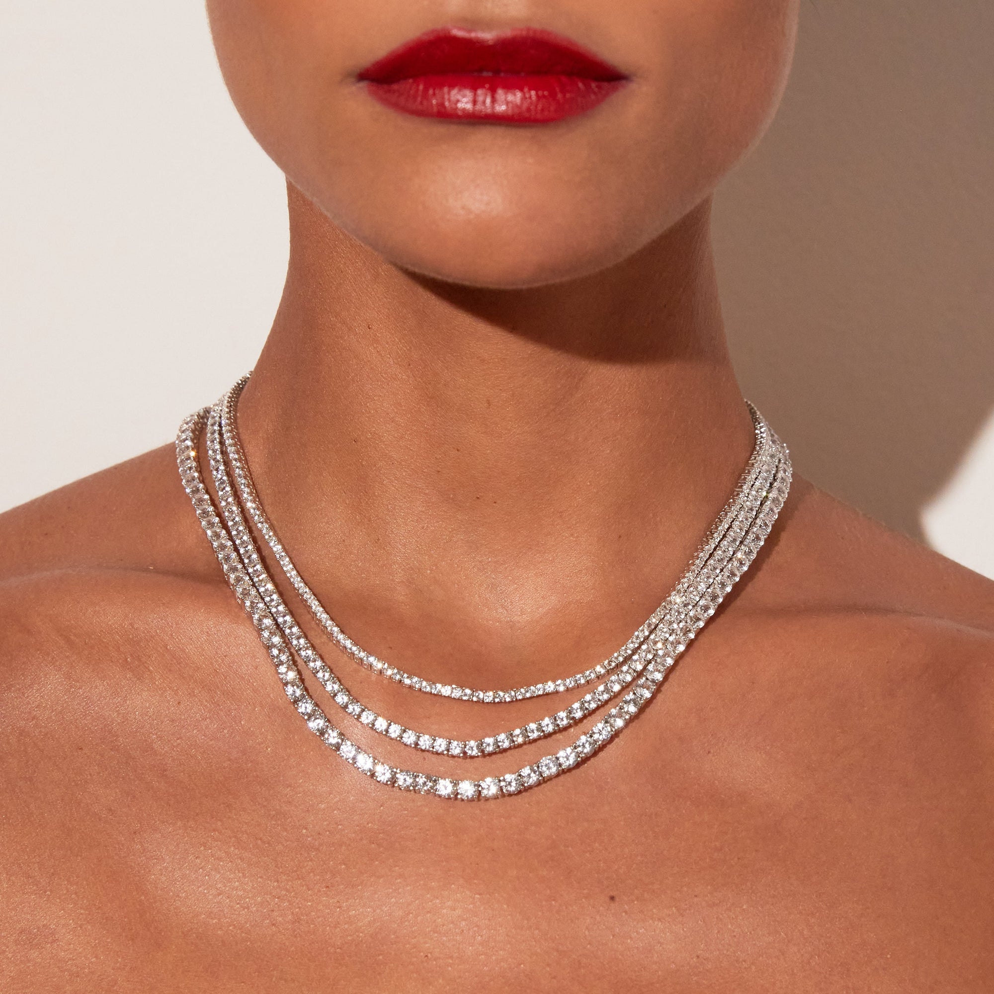 Close-up of a woman's neck and lower face featuring bright red lipstick, wearing three layered white sapphire tennis necklaces against a warm neutral background