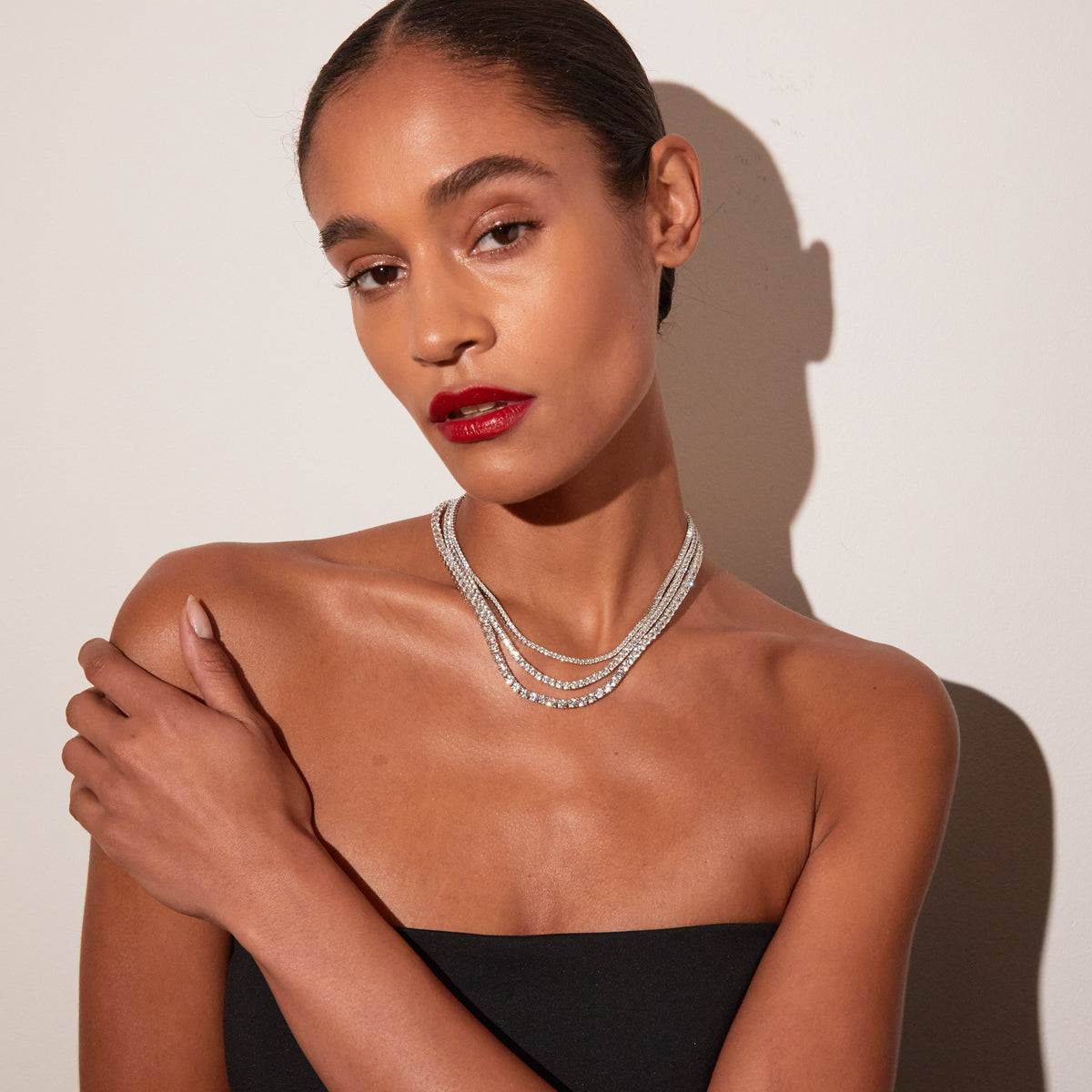Model. Portrait of a woman looking directly at the camera, wearing a black strapless top and bright red lipstick, with her hand gently resting on her bare shoulder. She is wearing three layered white sapphire tennis necklaces against a warm neutral background with a distinct shadow.