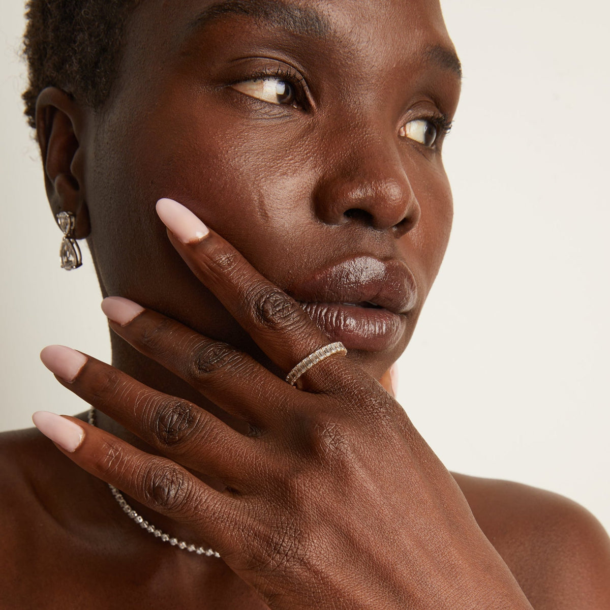 Model. Close-up of a woman's face with hand on cheek, wearing earrings and a ring, against a neutral background
