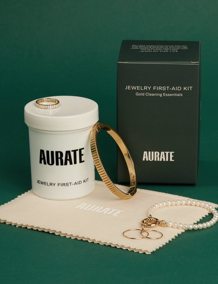 Flatlay Model A closed jar and the black packaging box of the Aurate Jewelry First-Aid Kit displayed on a green background with a branded polishing cloth, a gold bangle, a pearl bracelet, and gold rings.