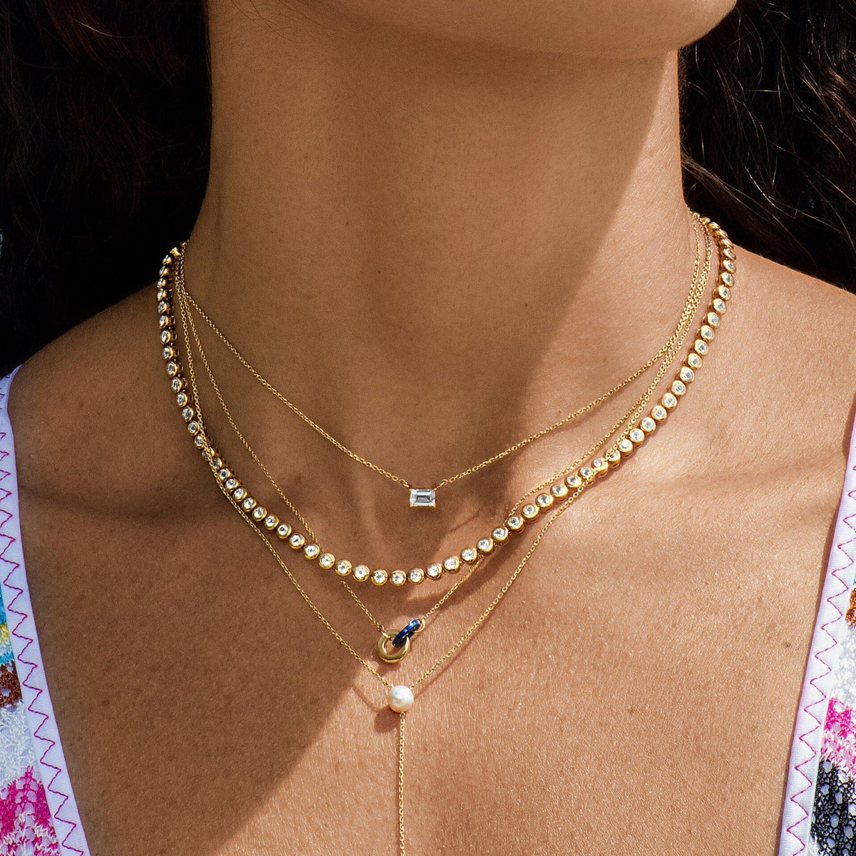 Lifestyle Model Close-up of a woman wearing a white top with pink trim and three layered necklaces, including a delicate chain with a small rectangular gemstone pendant, a diamond tennis necklace, and a delicate chain with a round blue gemstone and pearl drop pendant.