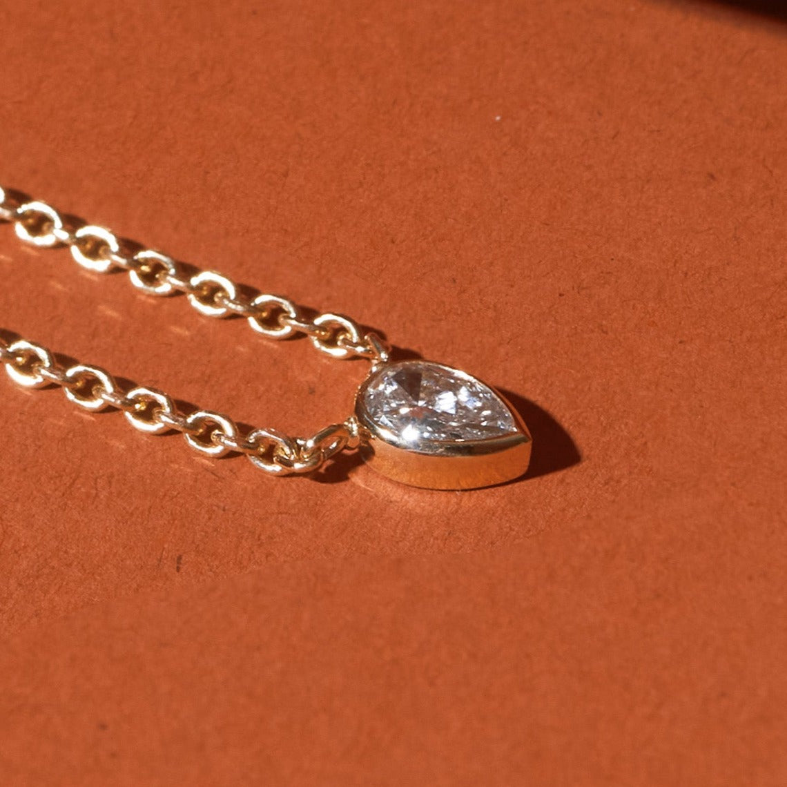 Flatlay Model Close-up of a delicate gold chain necklace featuring a pear-shaped, bezel-set diamond pendant, resting on a solid orange background and casting a distinct shadow.