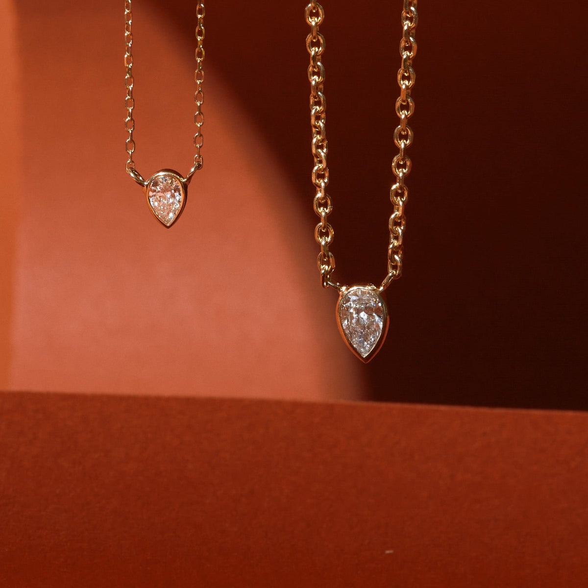 Flatlay Model Close-up of two delicate gold necklaces, each featuring a pear-shaped, bezel-set diamond pendant (one slightly larger than the other), suspended against a two-toned dark red and orange background.