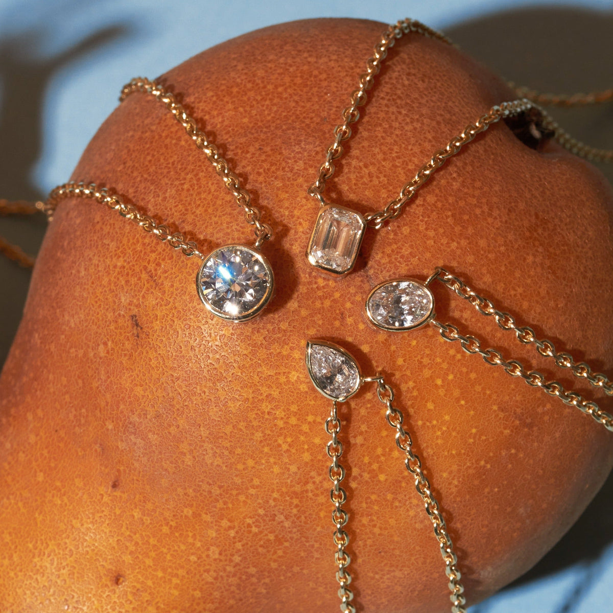 Flatlay Model Close-up of four delicate gold chain necklaces draped over a large, textured orange fruit, each featuring a different bezel-set diamond pendant: round, emerald-cut, oval, and pear-shaped.