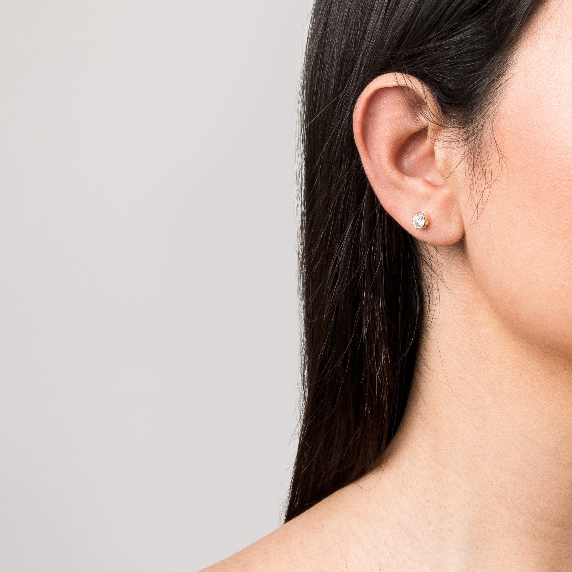 Model Close-up of a woman's profile and ear against a light background, wearing a round, bezel-set diamond stud earring. Her dark hair is tucked neatly behind her ear.