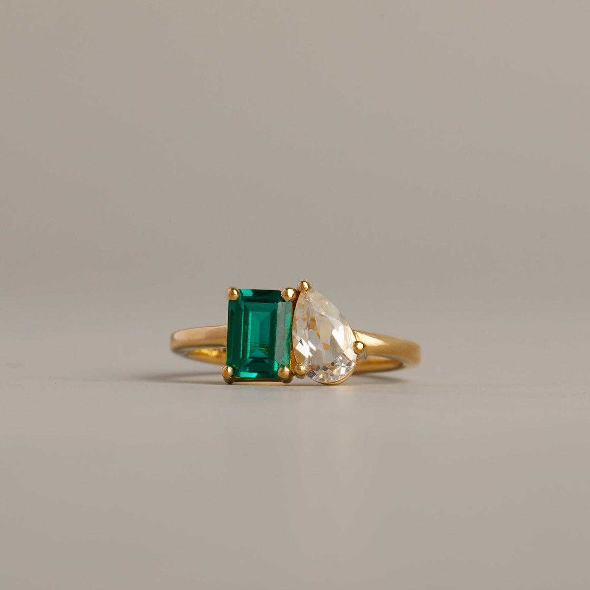 Model. Gold ring with green gemstone and heart-shaped clear gemstone on a beige background