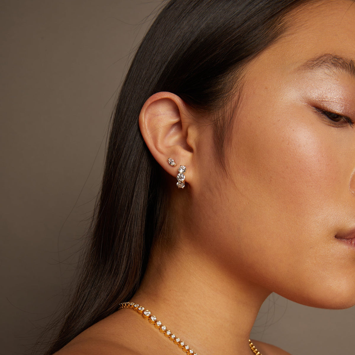 Model. Close-up of a woman wearing a gold necklace and diamond earrings against a neutral background