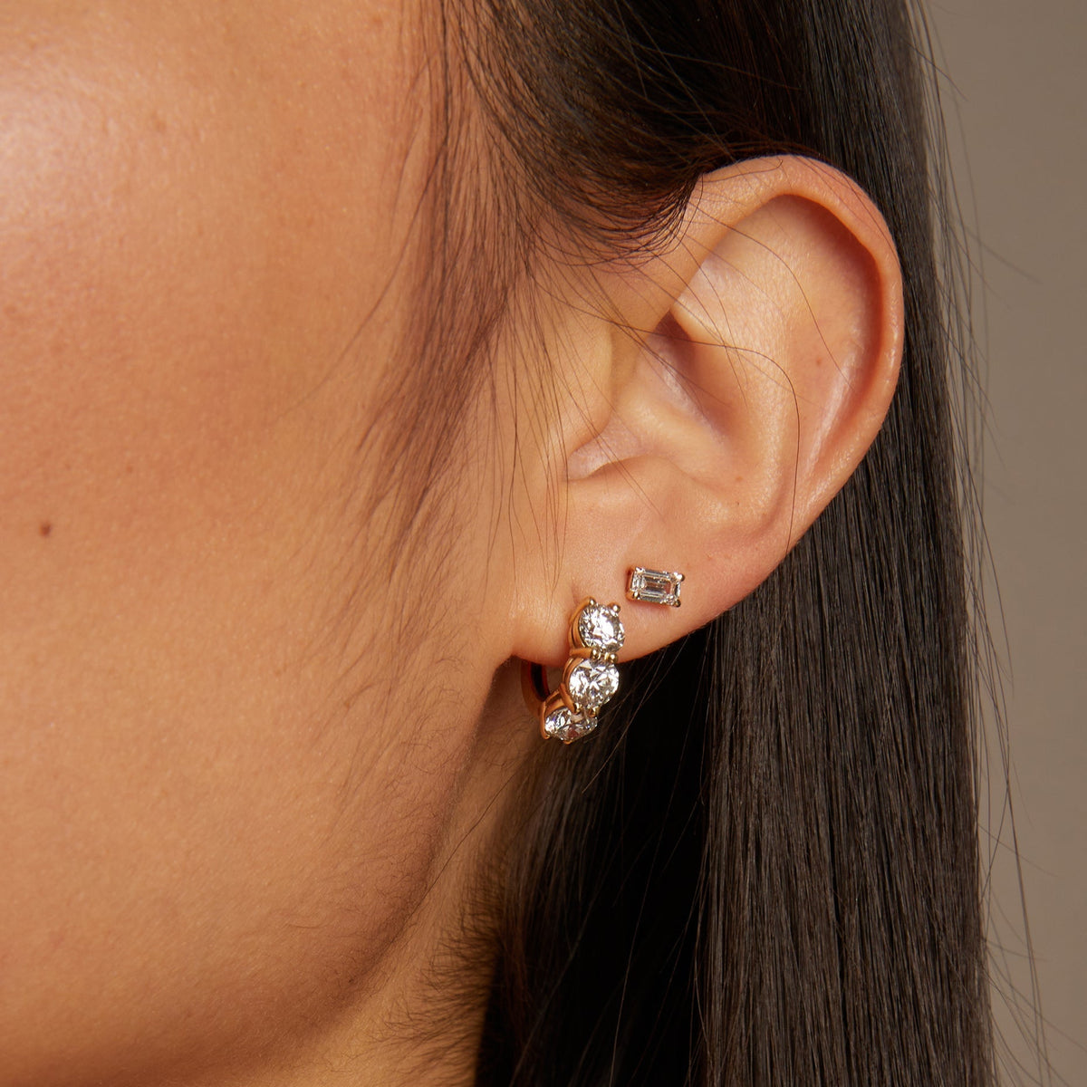 Model. Close-up of an ear wearing gold earrings with a neutral background