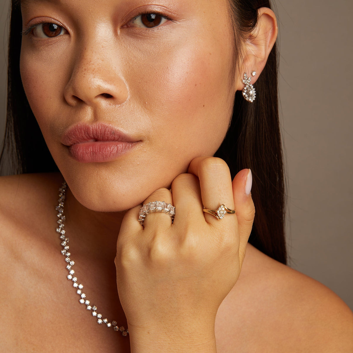 Model. Woman wearing jewelry including earrings, a necklace, and rings against a neutral background