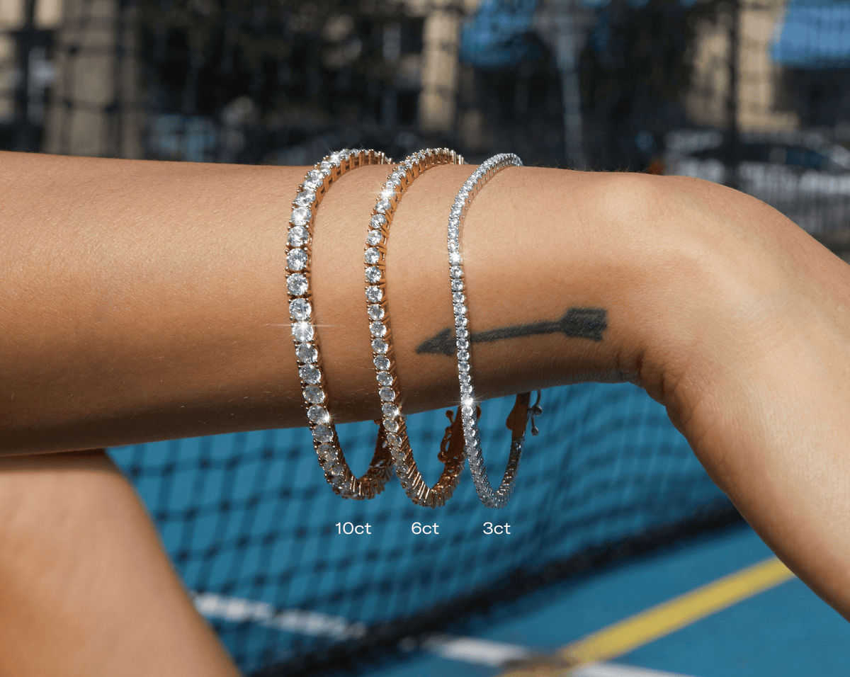Lifestyle Model Close-up of an arm with an arrow tattoo wearing three stacked white sapphire tennis bracelets against a blue tennis court background, with text overlays labeling them '10ct', '6ct', and '3ct'