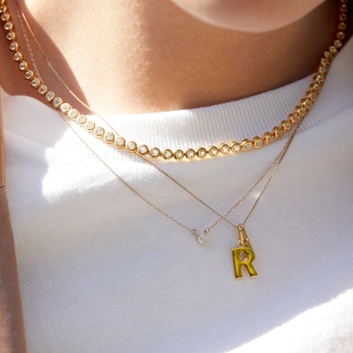 Lifestyle Model Close-up of a woman wearing a white top and three layered necklaces, including a diamond tennis necklace, a delicate chain with a single round gemstone pendant, and a delicate chain with a gold 'R' initial pendant.