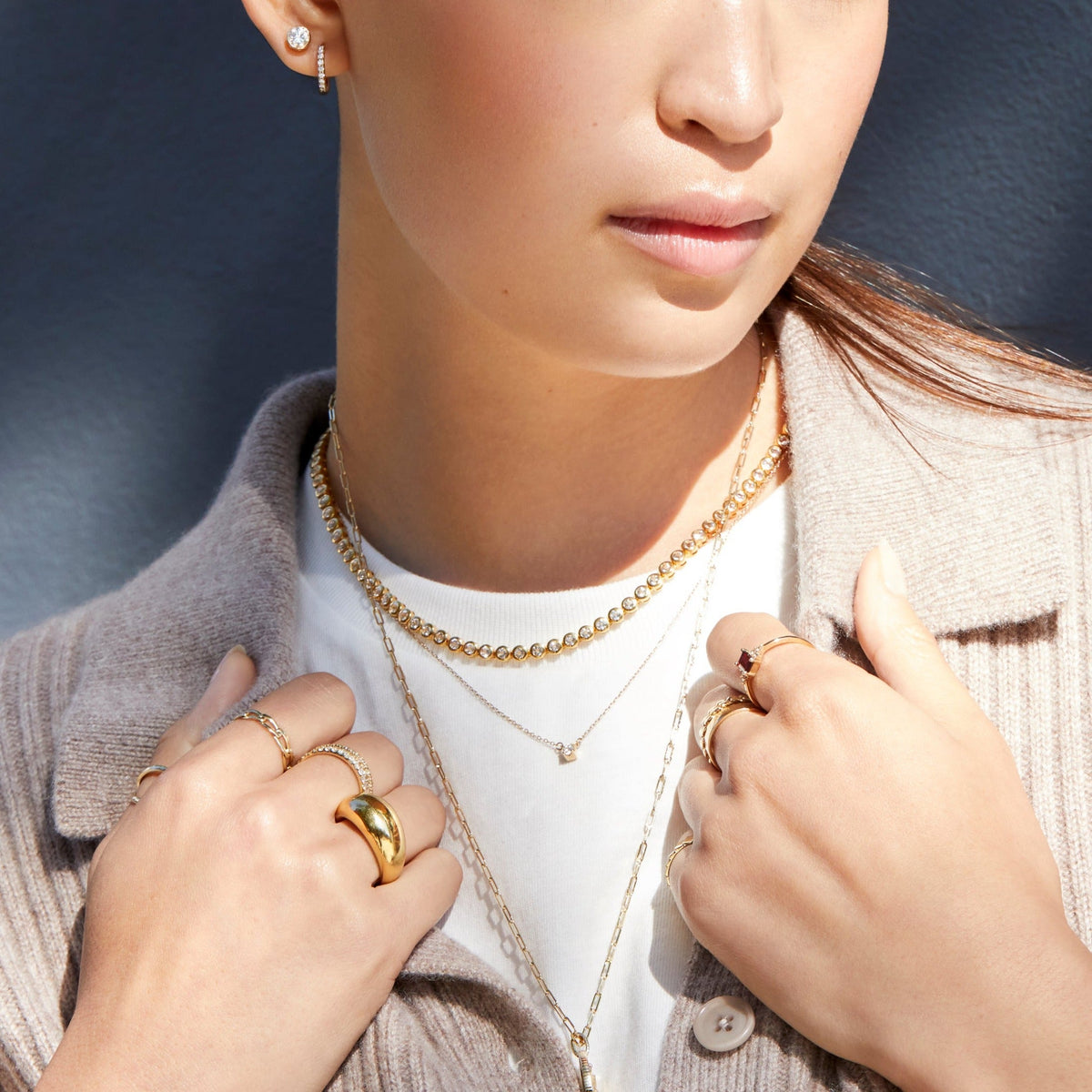 Lifestyle Model Close-up of a woman wearing a white top and a beige cardigan, accessorized with three layered necklaces, including a diamond tennis necklace and a delicate chain with a small gemstone pendant. Her hands are resting near her chest, showcasing multiple gold and diamond rings, including a thick gold dome ring and a ring with a dark red gemstone.