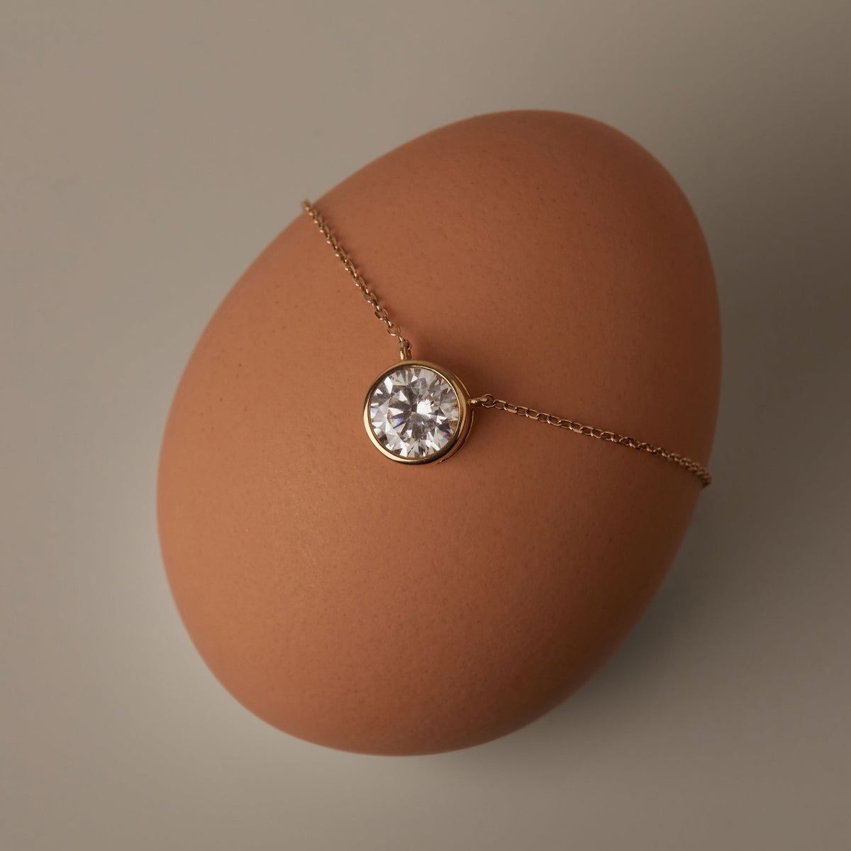 Flatlay Model Close-up of a delicate gold chain necklace featuring a round, bezel-set diamond pendant, resting on a smooth, light brown egg against a dark background.